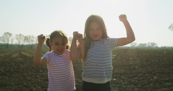 Authentic shot of two happy little girls are having fun to show their strength on a countryside green fields in a sunny day. Concept: happiness,fun, childhood, nature, authenticity, healthy lifestyle