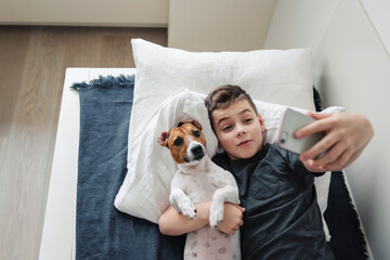 A young laughing boy lies on a bed next to a cute Jack Russell Terrier dog and takes a selfie.