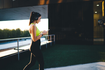 Slim sportswoman in active wear watching streams video on smartphone walking on training outdoors, charming fitness woman blogger holding cellular technology and connecting to 4g for web networking
