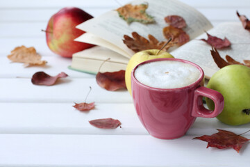 red cup with cappuccino with apples and an open book in fallen leaves. read warming stories