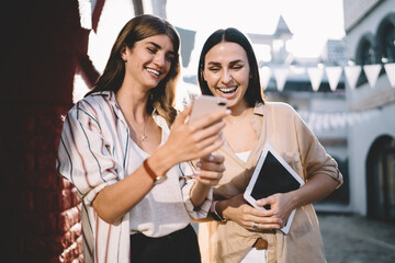 Happy female fellows discussing funny content during mobile social networking at urban setting, cheerful youthful women watching positive video vlog and smiling during friendly meeting in city