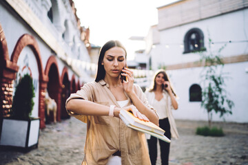 Fototapeta premium Caucasian female with textbook checking smartwatch time during international smartphone conversation, journalist with notepad looking at wrist while walking to meeting discussing info while calling