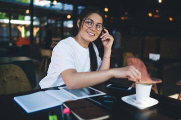 Cheerful caucasian millennial female in eyewear enjoying coffee break in cafe using modern digital device for communicating, smiling woman 20s blogger share multimedia content on web page via tablet