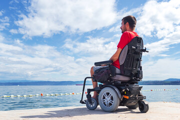 Man with muscular dystrophy on electric wheelchair outdoors looking at sea.
