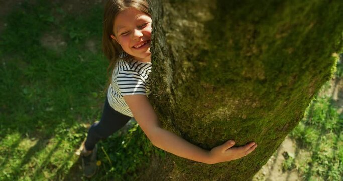 Authentic shot of a happy little girl is hugging a tree trunk and smiling in camera on a sunny day. Concept: love for nature, connection with nature, happiness, childhood, ecology, deforestation