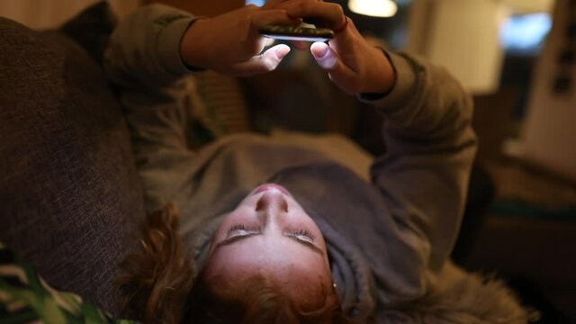 junge Frau liegt entspannt zuhause auf dem Sofa und schreibt kopf&uuml;ber auf ihrem Smartphone