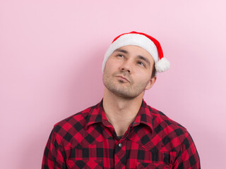 Emotions on the face, pensive, reflection, plan, idea. A man in a plaid rabbit and a Christmas red hat, on a pink background, copy space.