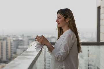 Happy young woman thinking and looking away with cup of tea or coffee with blurred city background....