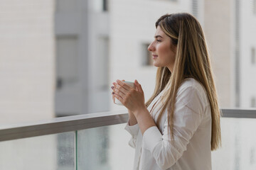 Young woman thinking and looking away with cup of tea or coffee with blurred city background. Girl...