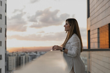 Young pretty woman dressed on white shirt looking at city from balcony, enjoying the sunset at terrace. Shot of beautiful young woman standing on the balcony and looking at the view. Relax, smiling.