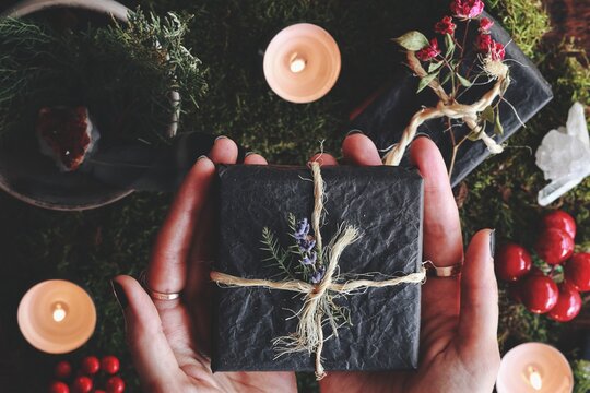 Yule winter solstice (Christmas) themed flat lay of female hands holding a black gift box wrapped in yarn and flowers in her hands. Forest moss, evergreens, burning lit candles, presents, red berries