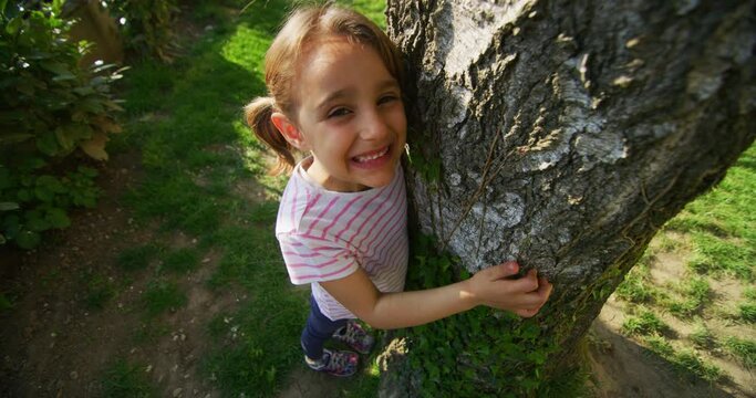 Authentic shot of a happy little girl is touching a tree trunk and smiling in camera on a sunny day. Concept: love for nature, connection with nature, happiness, childhood, ecology, deforestation