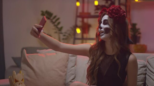 Slowmo PAN shot of happy young woman wearing flower crown and skull makeup sitting at couch at Halloween house party and taking selfie