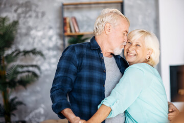 Happy senior wife and husband hugging at home, a man is kissing woman. Romantic middle-age couple has fun, dancing and laughing