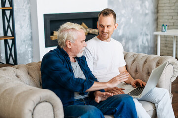 Cheerful two generation men family old senior father and smiling young adult son talking sitting on the comfortable sofa at home, using laptop for websurfing, online shopping