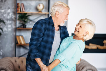 Portrait of modern romantic elderly couple is hugging, embracing gently, dancing at home. Senior spouses stand holding hands, looks to each other and laugh happily