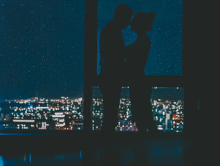 A loving couple are kissing on the background of the panoramic windows overlooking the city at night and the starry sky