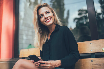 Caucasian cheerful hipster girl with perfect blonde hair taking rest on bench in city enjoying free...