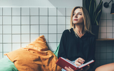 Contemplative hipster girl dressed in casual apparel holding education notepad for learning in loft...