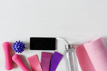 Dumbbells, fitness rubber bands, yoga mat, massage ball and smartphone on white wooden floor.