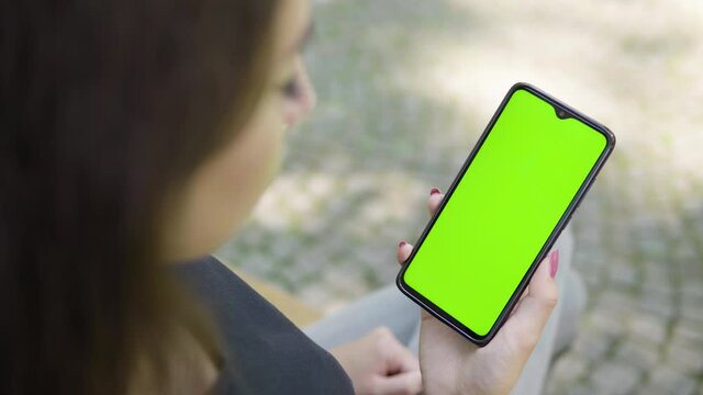 A black woman looks at a smartphone with green screen - vertical position - focused closeup from behind - she sits on a bench outside