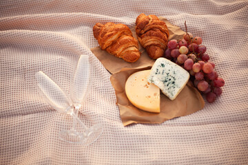 Romantic picnic for two at sunset. Two pieces of cheese, grapes, two croissants and two empty glasses are on the blanket. Close-up photo