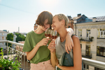 Two cheerful girls holding glass of wine, hugging while having party, celebrating anniversary on the balcony. Young lesbian couple spending their day together