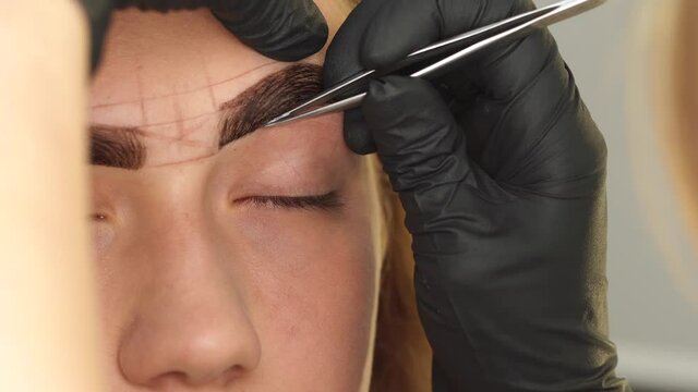 Professional styling, correction and lamination procedures of female eyebrows in beauty salon. Closeup view of client face and stylist's hands working in gloves. Master removes hair using tweezers.
