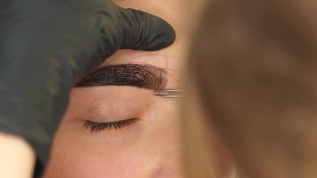 Professional styling, correction and lamination procedures of female eyebrows in beauty salon. Closeup view of client face and stylist's hands working in gloves. Master plucking hair using tweezers.