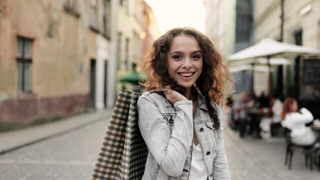 Beautiful fashionable model with curly hair holding shopping bags on her shoulder, looking at the camera and smiling. Portrait of young beautiful woman. Female nature beauty. Slow motion