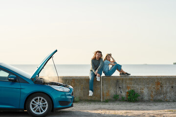 Outdoor shot of couple of travelers sitting near the broken car with open hood and smoking engine, man and woman looking tired, frustrated while waiting for assistance