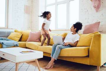 Playful daughter bouncing on sofa and looking at mother