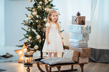 Fototapeta na wymiar a beautiful little girl in a white dress puts gifts on a sled near the Christmas tree. happy new year. retro-style. interior decoration for a holiday.