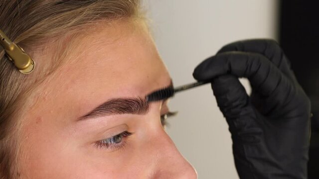 Professional styling, correction and lamination procedures of female eyebrows in beauty salon. Closeup view of client face and stylist's hands working in black gloves.