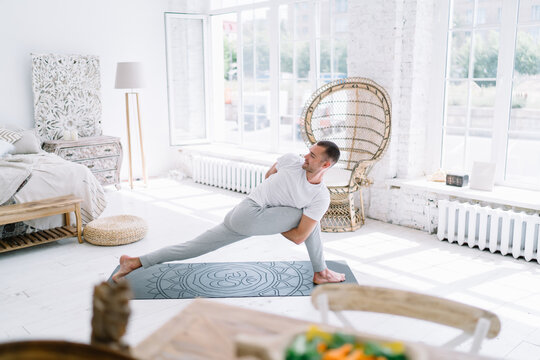 Smiling caucasian hipster guy in casual wear stretching muscle and keeping body strength on training, positive male standing in asana during morning yoga practice in clean modern interior home