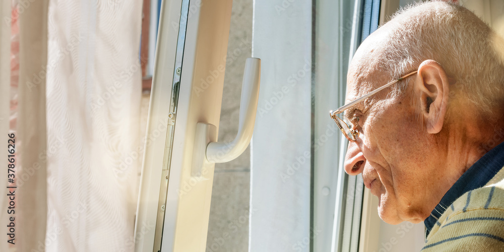 Wall mural bald old man stands at window looking outside closeup