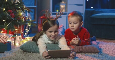 Caucasian small happy children, sister and brother, lying on the floor in the x-mas decorated dark room and watching cartoons on the tablet computer.
