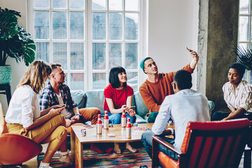 Obraz premium Caucasian hipster guy using front camera of cellphone for making selfie content during gather meeting with best friends, group of millennials spending weekend for poker wagering and drinks talking