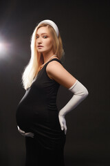 An elegant pregnant woman in a black evening dress and long white gloves posing with the sideways at the black background