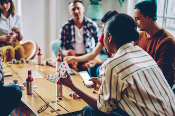 Back view of African American hipster girl with poker cards using mock up smartphone gadget with blank screen for casino betting advertising, female player chating via mobile during friendly gathering