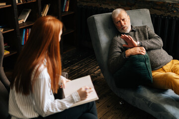 Professional red-haired young woman psychologist consulting mature man patient during psychological consultation. Lady taking notes in notebook, supporting and consoling depressed older male