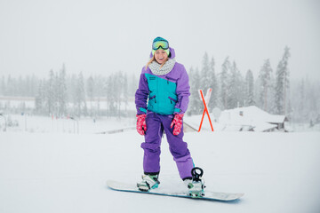 A young beautiful girl in sportswear and equipment stands in the winter on a mountain slope on a snowboard. Snowboarder laughs on a background of snow.