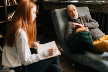 Serious red-haired young woman psychologist taking notes in notebook, supporting and consoling depressed older man patient during psychotherapy session.