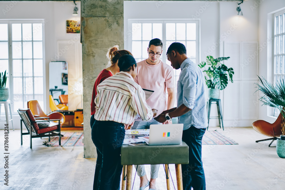 Poster multicultural people analyzing paper sketches standing at one table in modern office interior, skill