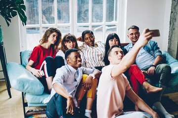 Diverse millennial people relaxing on comfortable sofa spending leisure time for making photo via smartphone camera, youthful friends taking selfie photo for sharing media content to website