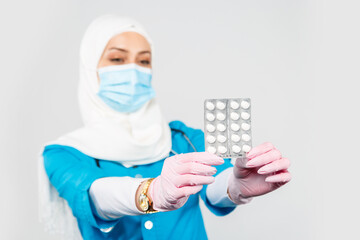 friendly Muslim doctor or nurse in a hijab, mask, gloves offering a pill to the patient on a gray background