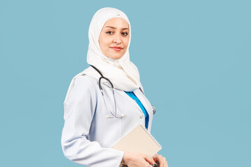 friendly, Muslim doctor or nurse woman in a hijab with a stethoscope writes a prescription on pills on a blue background.