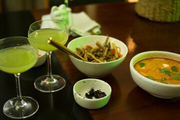 White dragon cocktail glasses with a plate of tom yam, crickets and pork ears salad on a wooden table

