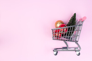 Small shopping cart with of festive balls and Christmas tree, pink background, horizontal, copy space