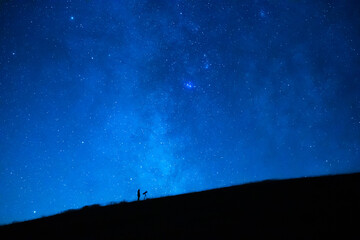 Astronomer observing the immensity of the universe and the stars. Small silhouette of an astronomy lover with a telescope observing the blue starry sky at night.